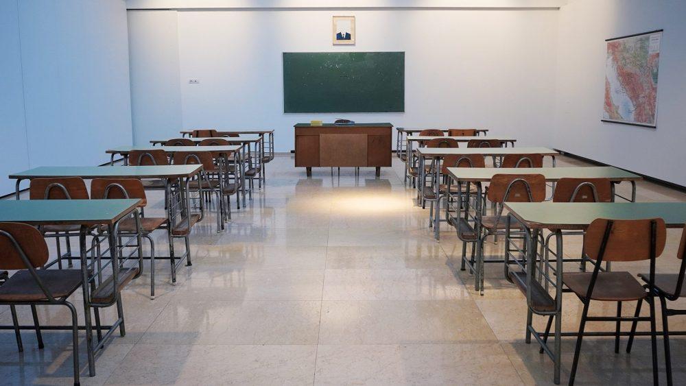 Foto: Leeres Klassenzimmer mit mehreren Tischen und Stühlen aus Holz in Reihen. An der hinteren Wand eine grüne Tafel, darunter ein brauner Schrank. Weiße Wände, helles Licht von oben. Rechts an der Wand ein Poster. Bildnachweis: Ivan Aleksic auf Unsplash.