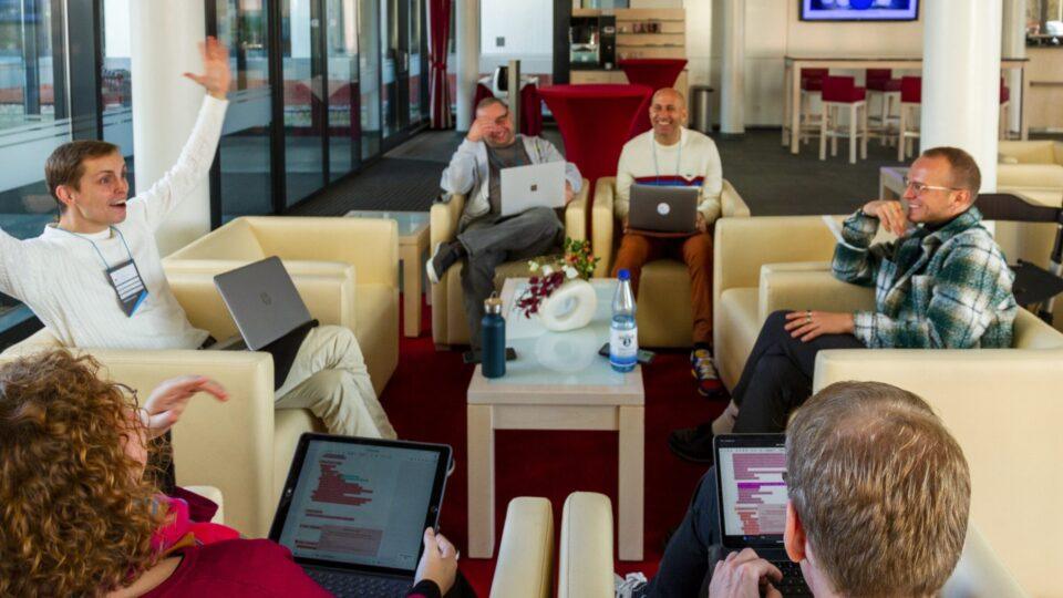 Mehrere auf Sesseln sitzende Menschen unterhalten sich angeregt. Alle haben aufgeklappte Laptops auf ihren Schößen stehen.