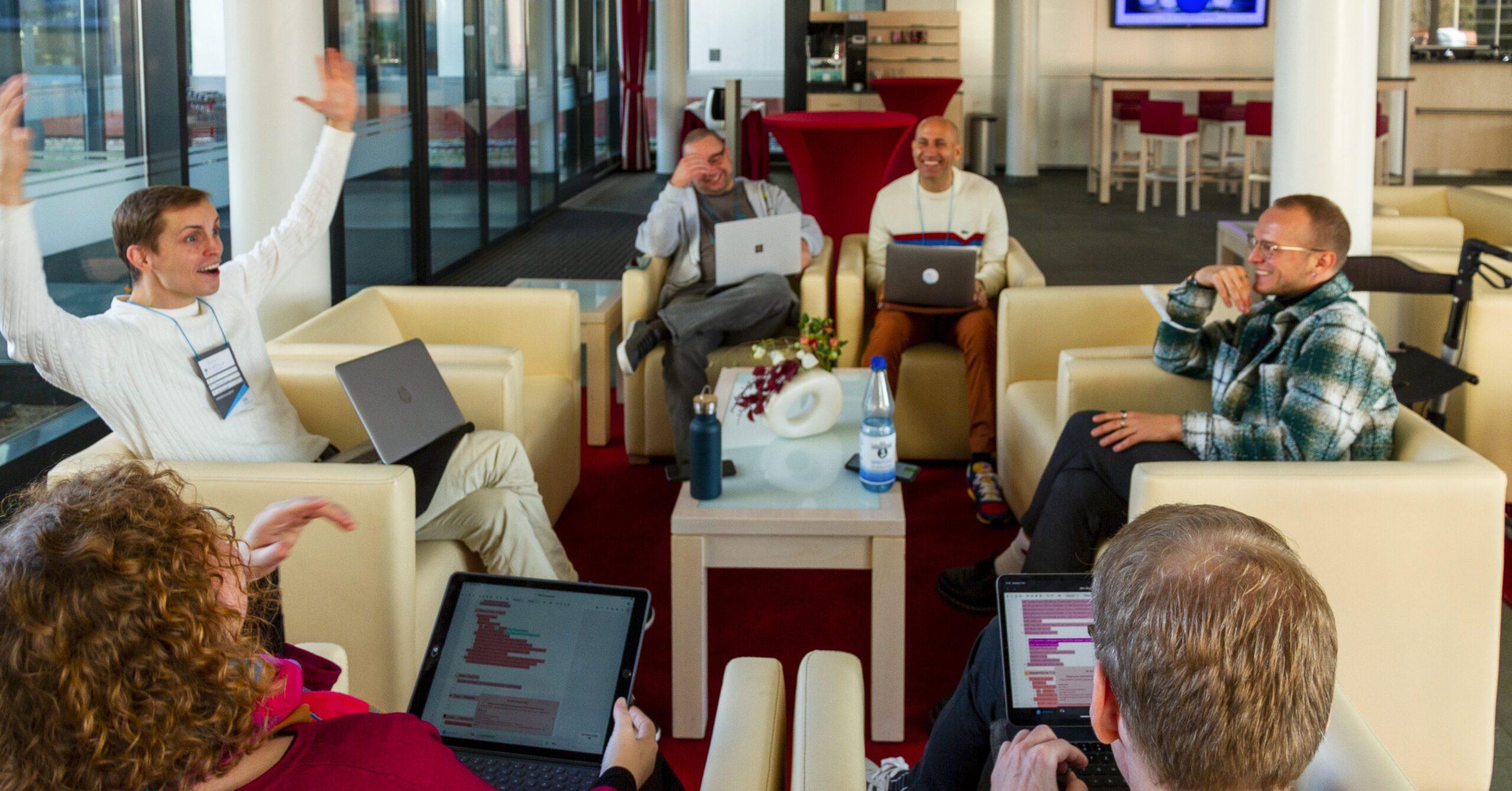 Foto: Mehrere auf Sesseln sitzende Menschen unterhalten sich angeregt. Alle haben aufgeklappte Laptops auf ihren Schößen stehen.