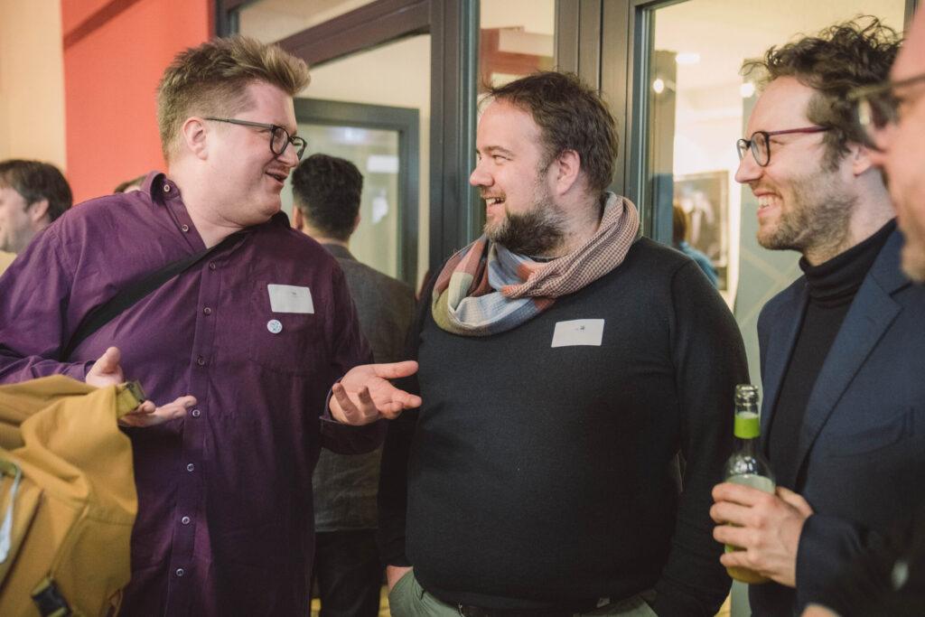 Foto: Drei Personen unterhalten sich und lachen in einer Indoor-Veranstaltung. Links eine Person mit Brille, kurzem hellbraunem Haar, dunkelrotem Hemd, Namensschildhalter, hält ein gelbes Getränk. Mitte eine Person mit Bart, dunkelbraunem Haar, schwarzem Sweatshirt, kariertem Schal, Namensschildhalter, hält ein grünes Getränk. Rechts eine Person mit Brille, lockigem dunkelbraunem Haar, dunklem Pullover, hält ein grünes Getränk. Im Hintergrund weitere Personen verschwommen.