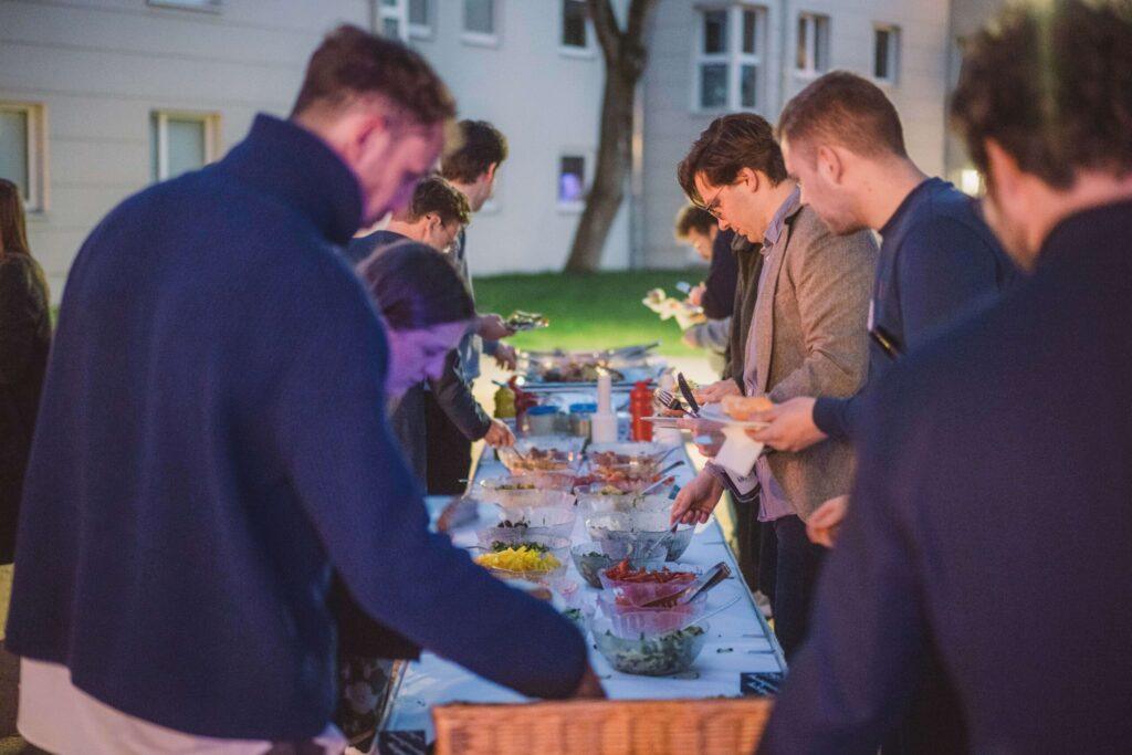 Foto: Abendliche Veranstaltung. Etwa 8-10 Personen stehen an einem langen Tisch mit verschiedenen Speisen und Getränken. Verschiedene bunte Schüsseln mit Salaten, Saucen und anderen Lebensmitteln. Im Hintergrund helles Gebäude mit Fenstern, Baum, grüner Rasen. Personen bedienen sich vom Buffet.