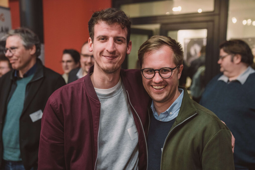 Foto: Zwei Männer im Vordergrund einer Veranstaltung stehen nebeneinander und schauen in die Kamera. Links ein Mann mit lockigem braunem Haar und Bart trägt eine bordeaux-rote Jacke über grauem Hemd und ein Namensschild. Rechts ein Mann mit hellbraunem Haar und Brille lächelt breit, trägt einen olivgrünen Pullover über hellblauem Hemd. Im Hintergrund weitere Personen bei einer Veranstaltung.