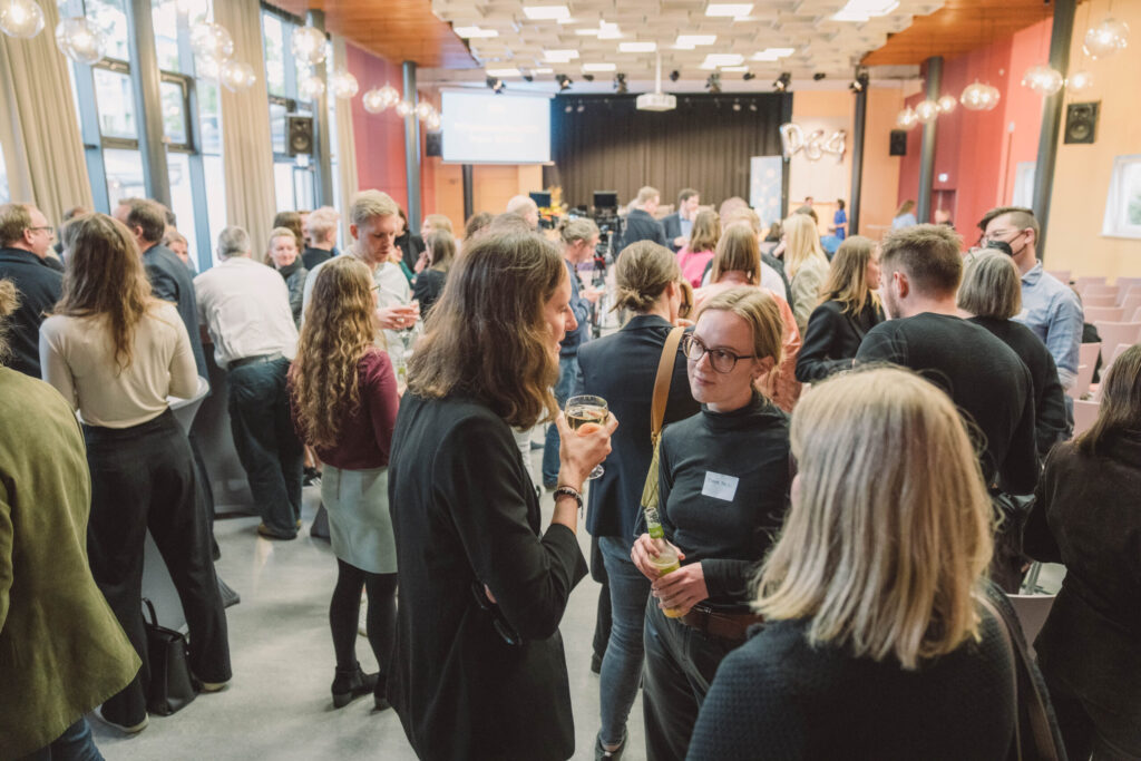 Foto: Viele Personen bei einer Veranstaltung in einem großen hellen Saal mit farbigen Wänden, modernen Deckenleuchten und großen Fenstern. Sie stehen durcheinander und unterhalten sich. Im Vordergrund: Person mit langen dunklen Haaren in schwarzer Kleidung, hält ein Glas Wasser. Daneben: Person mit Brille, schwarzem Rollkragenpullover und gelbem-braunem Schultergurt mit Badge. Im Hintergrund Bühne mit Vorhang, weitere Personen bei Gesprächen. Entspannte Networking-Atmosphäre.