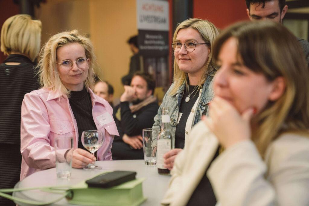 Foto: Mehrere Personen bei einer Veranstaltung an einem Tisch. Vorne links eine Frau mit blondem Haar und Brille trägt eine rosa Jacke über schwarzem Oberteil, hält ein Glas. Neben ihr eine Frau mit blondem Haar, Brille, grauem Cardigan und Perlenkette, trägt ein Namensschild-Band. Vorne rechts (unscharf) eine Frau mit dunkelbraunem Haar in beigem Oberteil. Auf dem Tisch Gläser. Im Hintergrund weitere Personen und ein schwarzes Banner.
