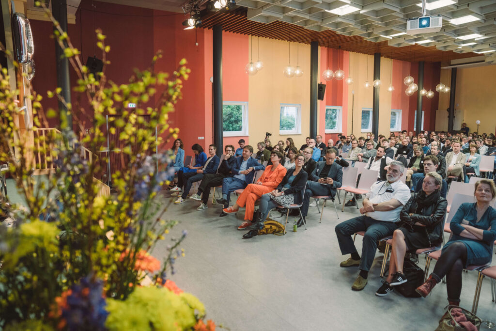 Foto: Veranstaltung in einer großen Halle mit farbigen Wänden. Etwa 100–120 Personen sitzen auf Stühlen und schauen nach vorne. Hängende Pendelleuchten in warmem Weiß, große Fenster. Im Vordergrund unscharfe Blüten mit gelben und roten/orangenen Blüten. Verschiedene Altersgruppen, diverse Kleidung.