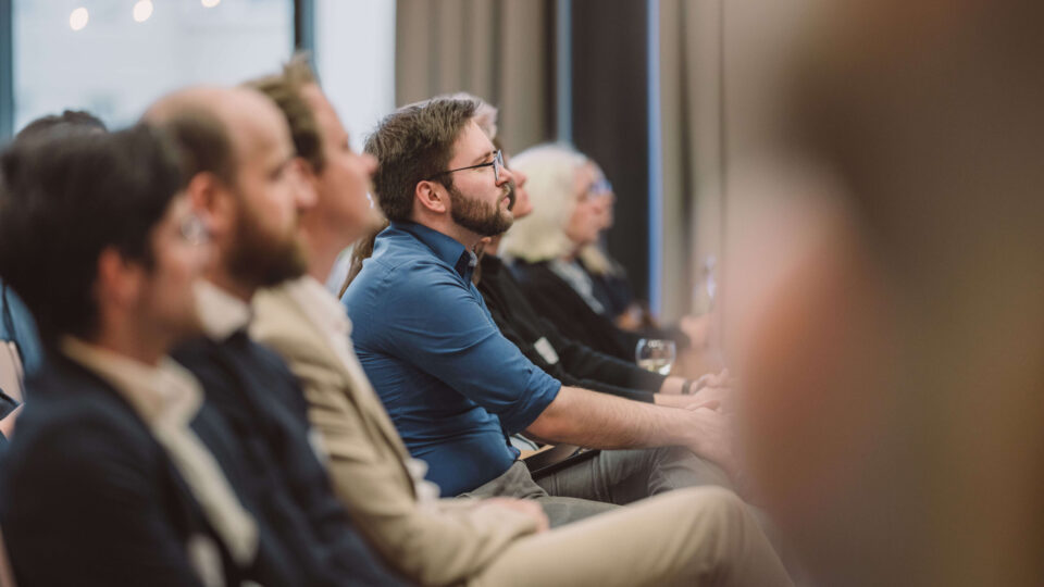 Foto: Menschen, die einer Veranstaltung zuhören. Ein Mann ist fokussiert, Menschen in Sitzreihen sind verschwommen erkennbar.