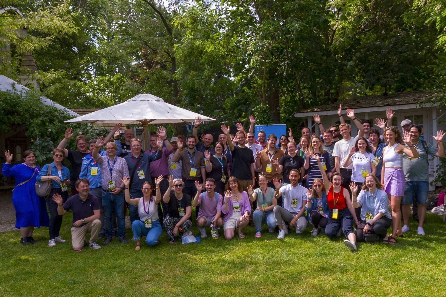 Foto: Gruppenfoto der Teilnehmer:innen des D64-Meetups auf der re:publica 23. Etwa 60–70 Personen auf einer Wiese winken in die Kamera. Viele tragen gelbe Namensschild-Bänder. Verschiedene Altersgruppen, diverse Kleidung. Im Hintergrund beige-grauer Schirm, Bäume und grüne Vegetation, Gebäude teilweise sichtbar. Sonniges Wetter.