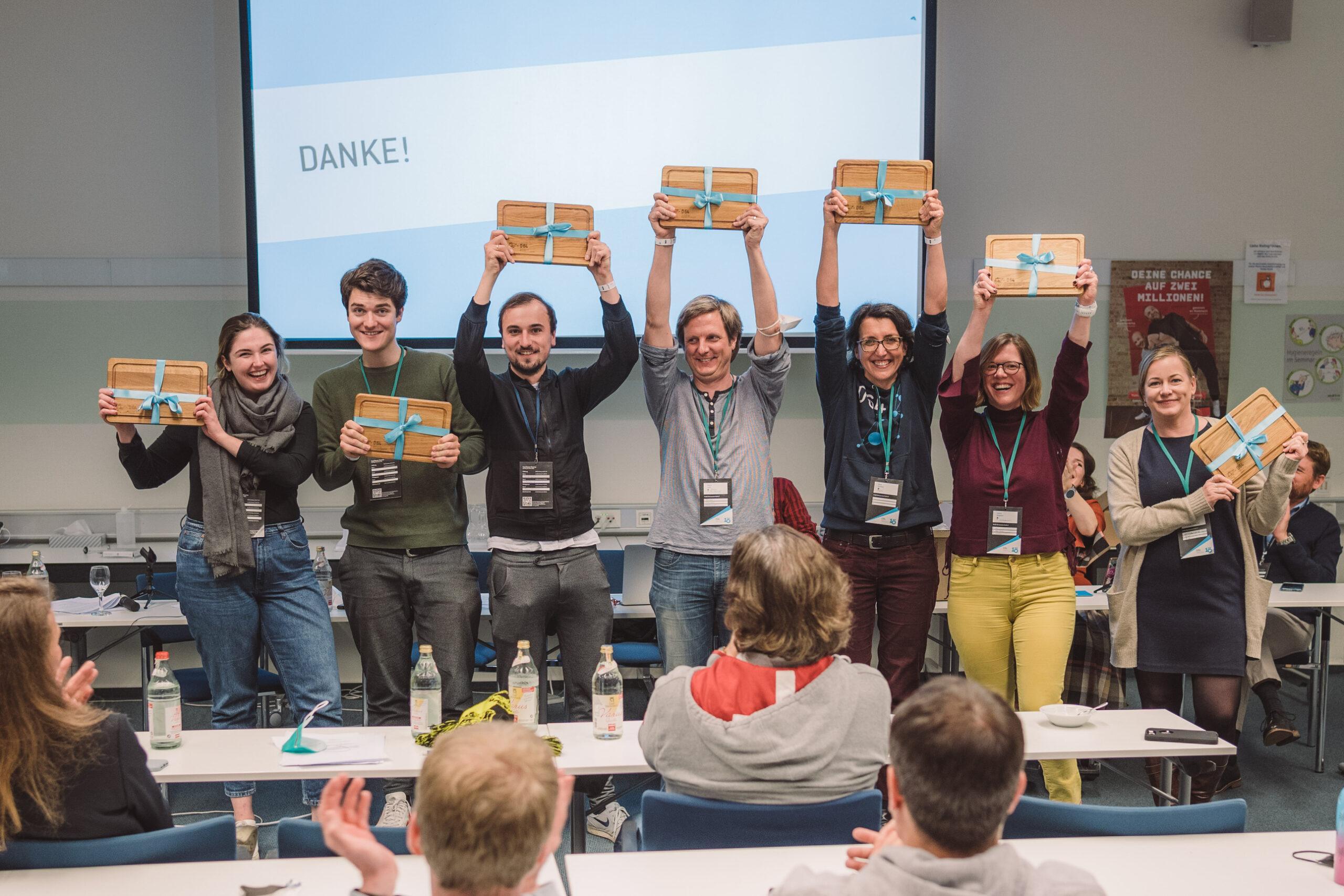 Foto: 7 Mitglieder des Tickerteams halten Frühstücksbrettchen mit hellblauer Schleife hoch. Hinter ihnen steht auf einer Leinwand: DANKE!