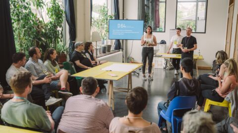 Eine Gruppe an sitzenden Menschen diskutiert in einem hellen, offenen Raum auf der re:publica 2023 mit drei D64-Mitgliedern, die vor einem Präsentationsmonitor stehen.