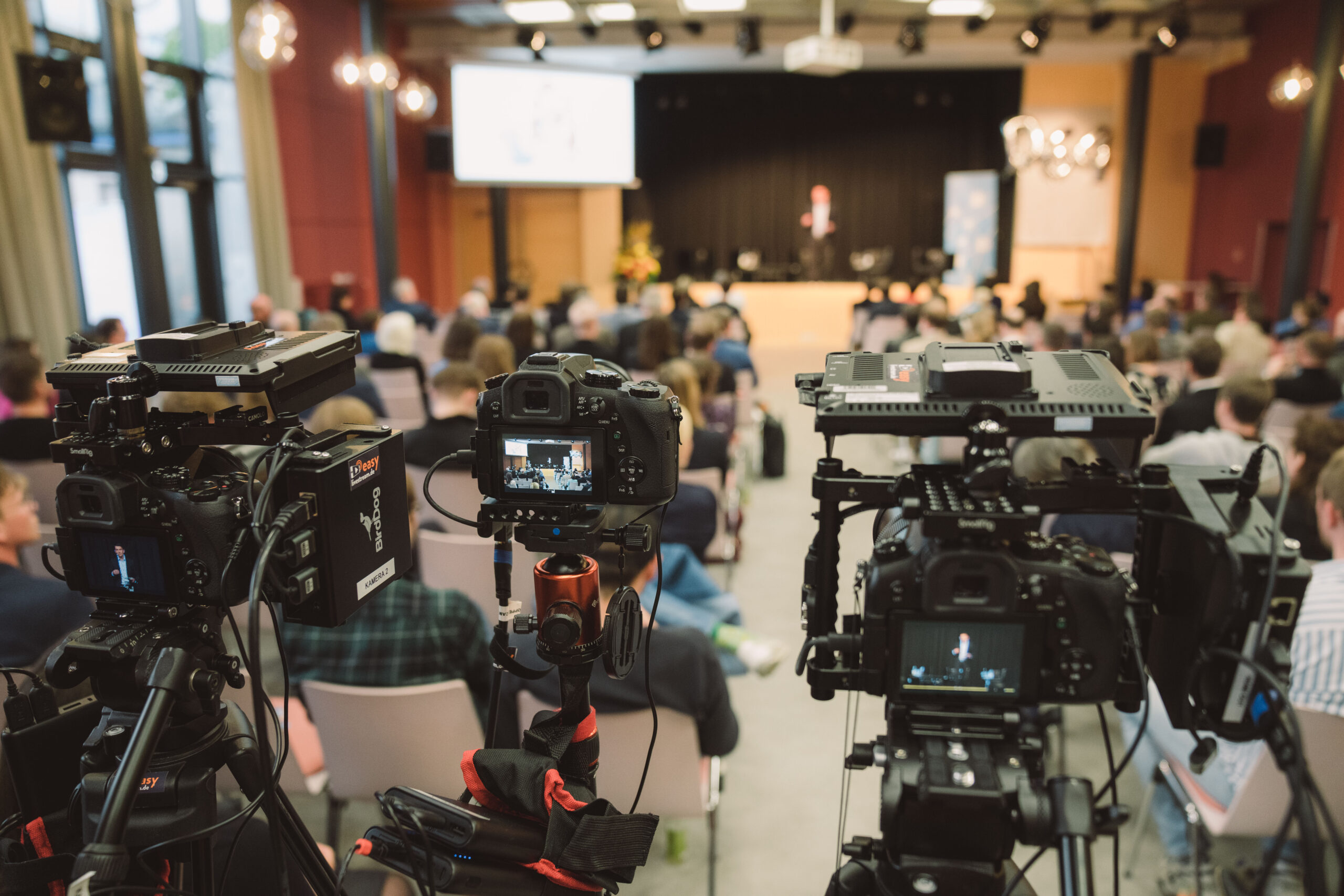 Drei Kameras inklusive Tonaufnahmegeräte filmen einen großen Saal mit einem Podium, welches im Hintergrund des Bildes zu erkennen ist. Auf dem Podium befindet sich ein Redner und vor ihm sitzen auf Stühlen, den Kameras abgewandt, viele Zuschauer:innen.