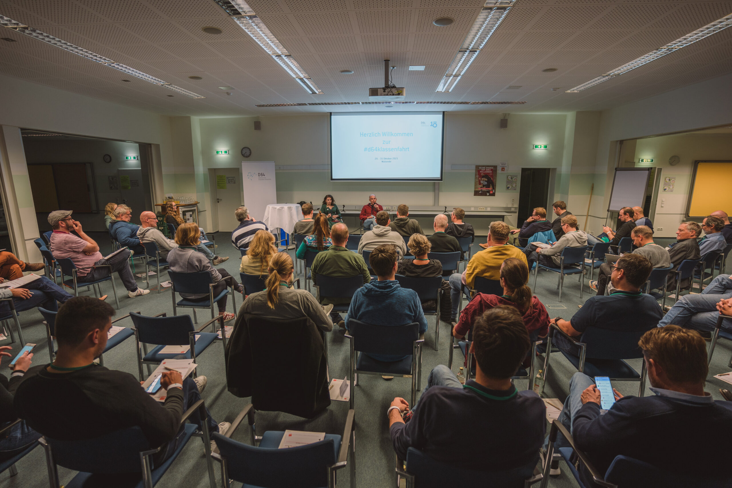 D64-Mitglieder sitzen in einem Konferenzraum auf Stühlen und diskutieren in einer moderierten Diskussion. Vorne vor den Reihen ist eine Leinwand aufgebaut, auf die Inhalte projiziert werden.