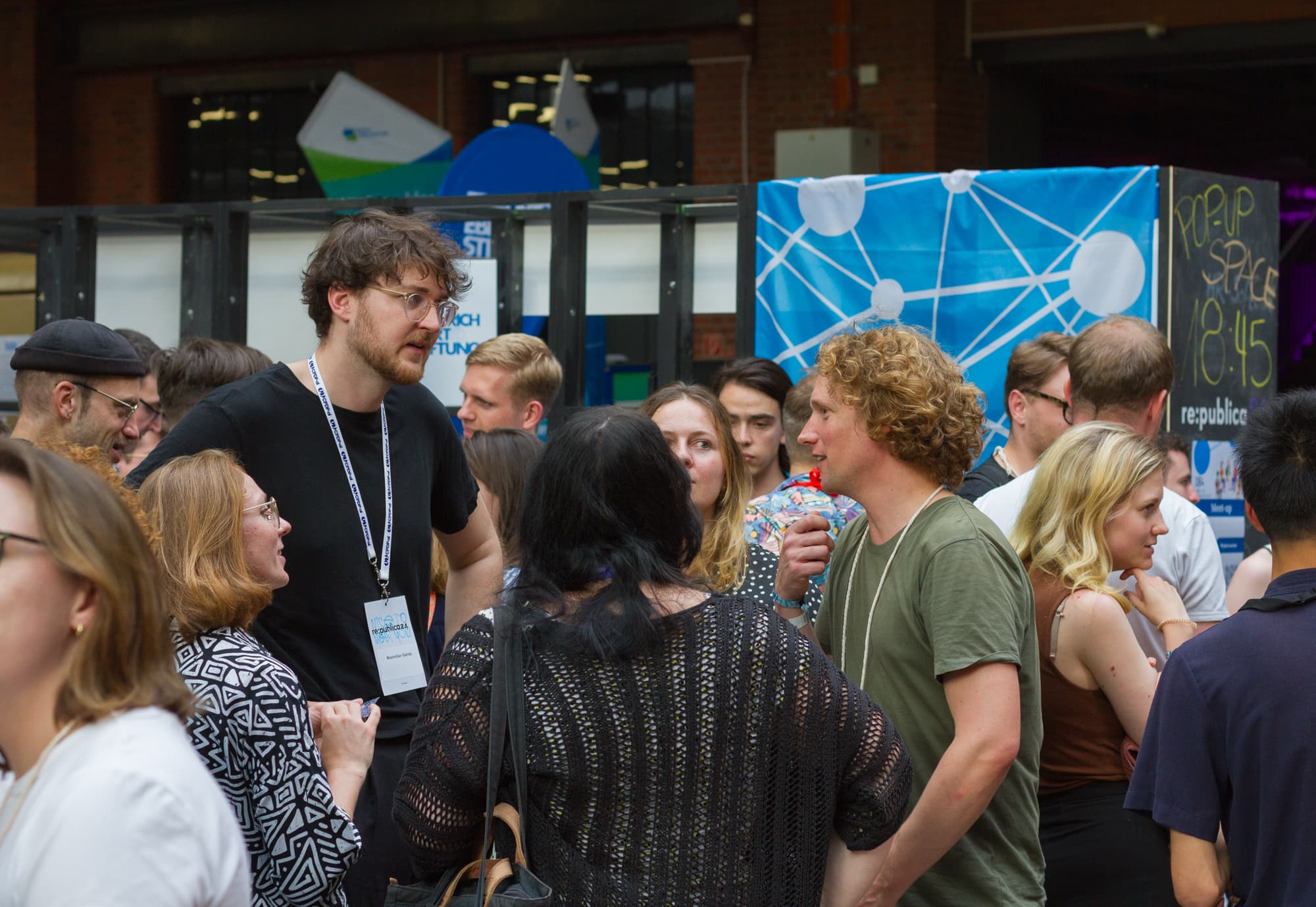 Foto: D64-Mitglieder und Interessierte im Gespräch. Es sind etwa 20 Personen zu sehen in einem Stand in der großen Halle der re:publica. Am Stand hängt ein D64-Banner. Bildnachweis: D64/Johann Lensing.