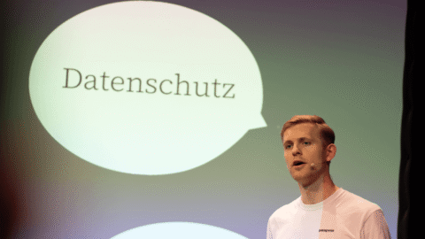 Foto: Torben Dzillak spricht bei der re:publica auf einer Bühne vor einer Projektionsfläche mit grün-violetter Beleuchtung. Die Projektion zeigt zwei Sprechblasen mit den Worten „Datenschutz" und „Sicherheit". Er trägt ein weißes T-Shirt und hält ein Gerät oder Buch. Im Vordergrund von links teilweise unscharfe Person. Bildnachweis: D64/Falko Ortloff.