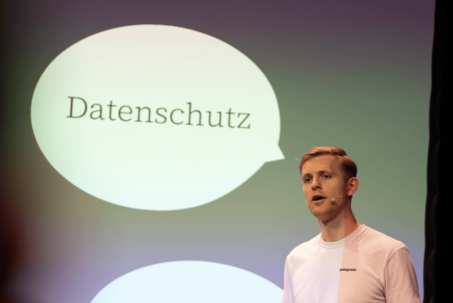 Foto: Torben Dzillak spricht bei der re:publica auf einer Bühne vor einer Projektionsfläche mit grün-violetter Beleuchtung. Die Projektion zeigt zwei Sprechblasen mit den Worten „Datenschutz" und „Sicherheit". Er trägt ein weißes T-Shirt und hält ein Gerät oder Buch. Im Vordergrund von links teilweise unscharfe Person. Bildnachweis: D64/Falko Ortloff.
