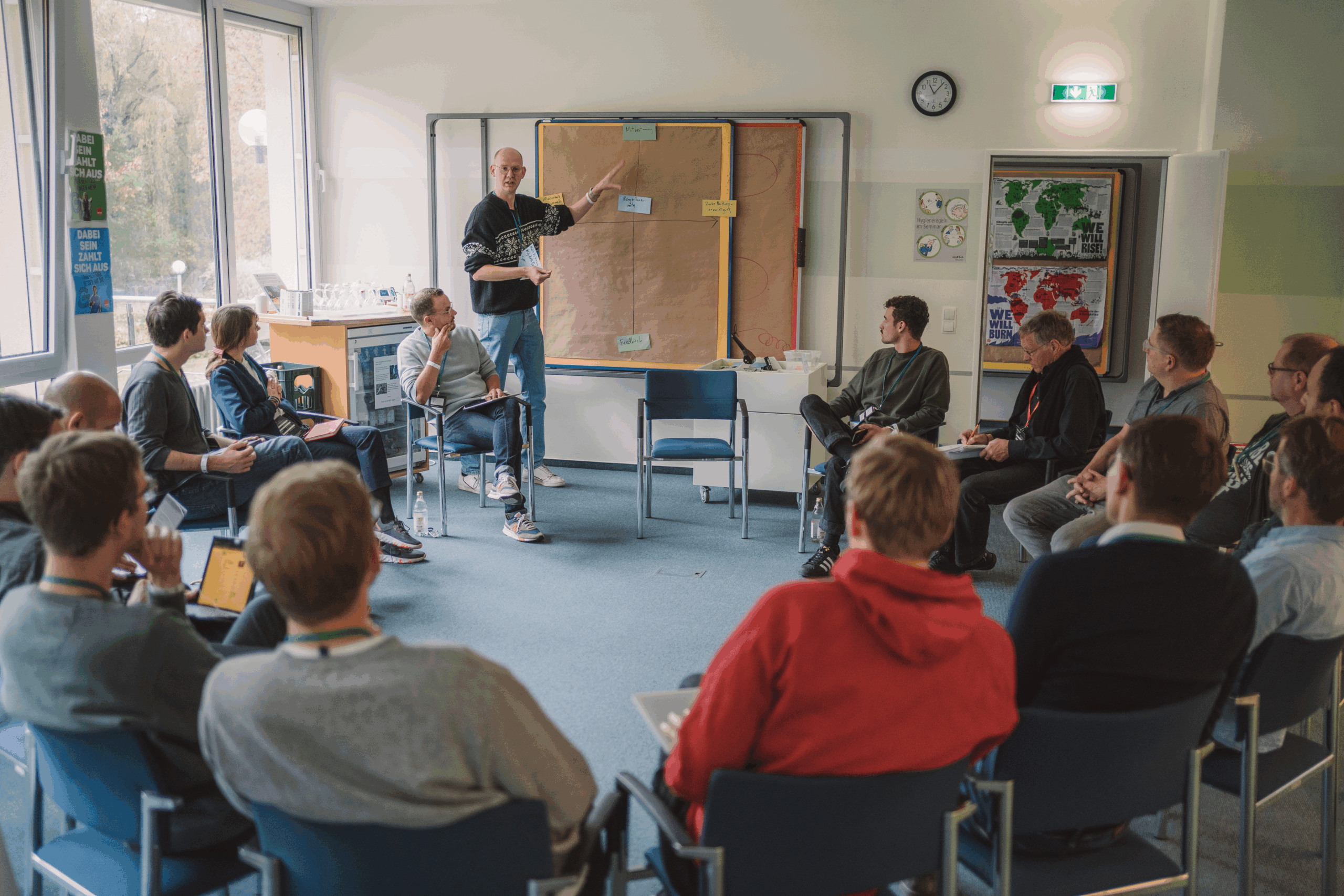 Foto: Workshop oder Diskussionsrunde. Etwa 15–20 Personen sitzen auf Stühlen im Kreis. Eine Person in der Mitte gestikuliert und spricht vor einer großen Plakattafel. Im Hintergrund Fenster, Plakate an den Wänden, Uhren. Bildnachweis: D64/Fionn Große.