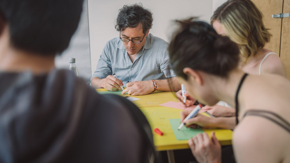 Foto: Workshop-Szene in einem hellen Raum. Mehrere Personen sitzen um einen gelben Tisch und schreiben mit farbigen Markern auf bunte Moderationskarten. Eine Person in der Mitte konzentriert sich auf die Tischfläche. Im Vordergrund von hinten zu sehen eine teilweise unscharfe Person.