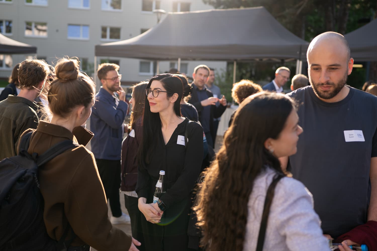 Foto: Networking-Szene bei einer D64-Veranstaltung. 20 Personen stehen in Zweiergruppen im Gespräch auf einem sonnigen Außengelände zusammen. Mehrere Teilnehmende tragen Namensschilder mit D64-Logo. Im Hintergrund sind Catering-Pavillons aufgebaut.