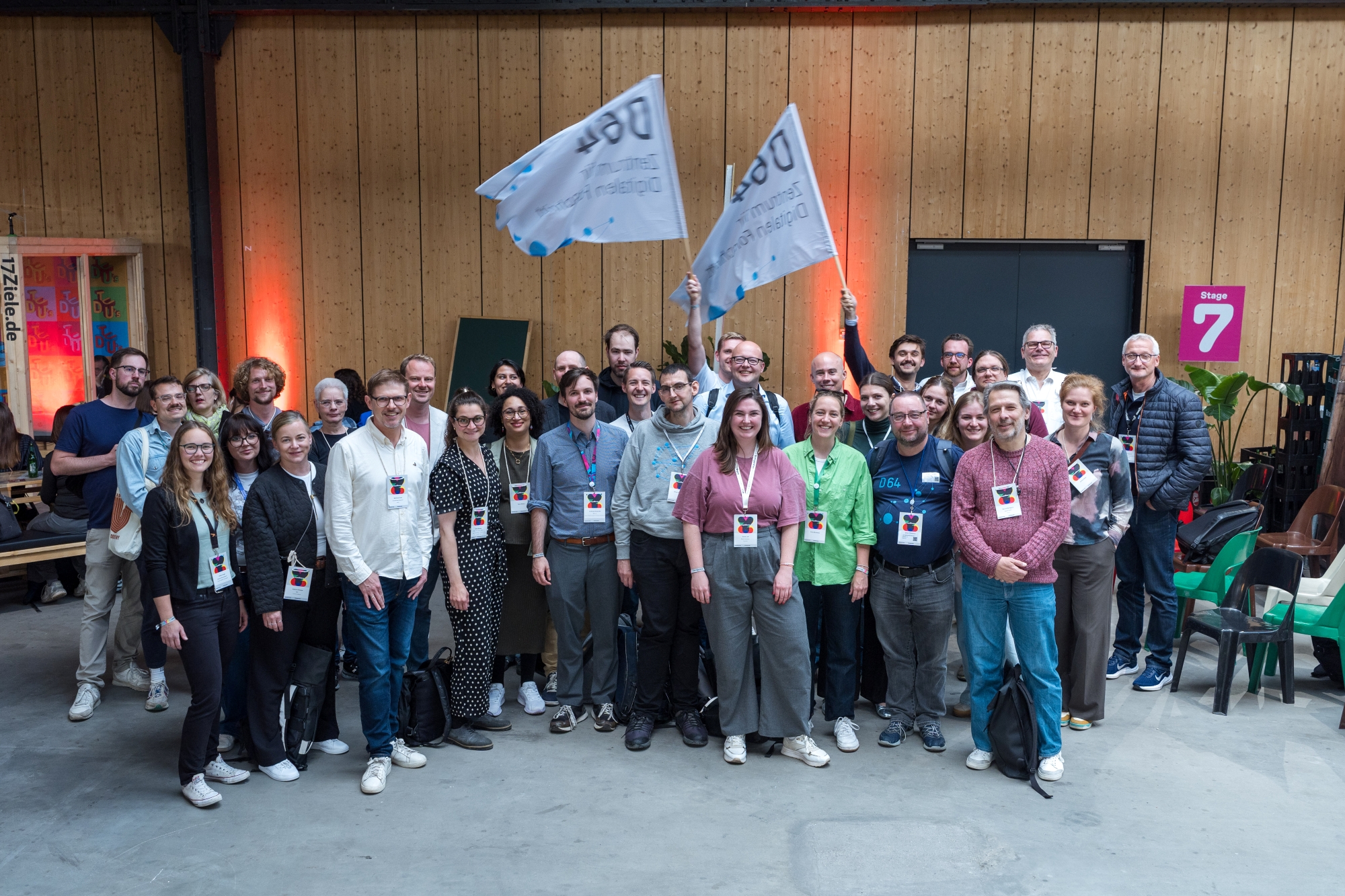 Foto: Gruppenfoto von etwa 40 Personen in einer Halle mit Holzwand. Alle tragen Namensschilder. Zwei Personen in der Mitte halten weiße Flaggen mit D64-Logo hoch. Rote und orangefarbene Beleuchtung im Hintergrund.