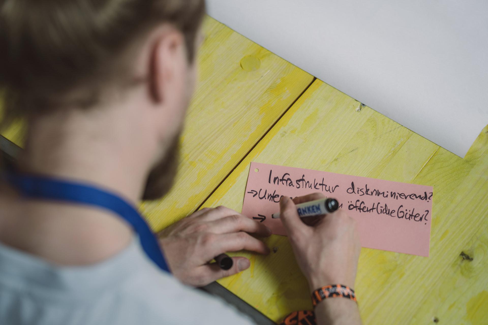 Ein Mensch schreibt auf eine Moderationskarte "Infrastruktur diskriminierend? .. öffentliche Güter?"