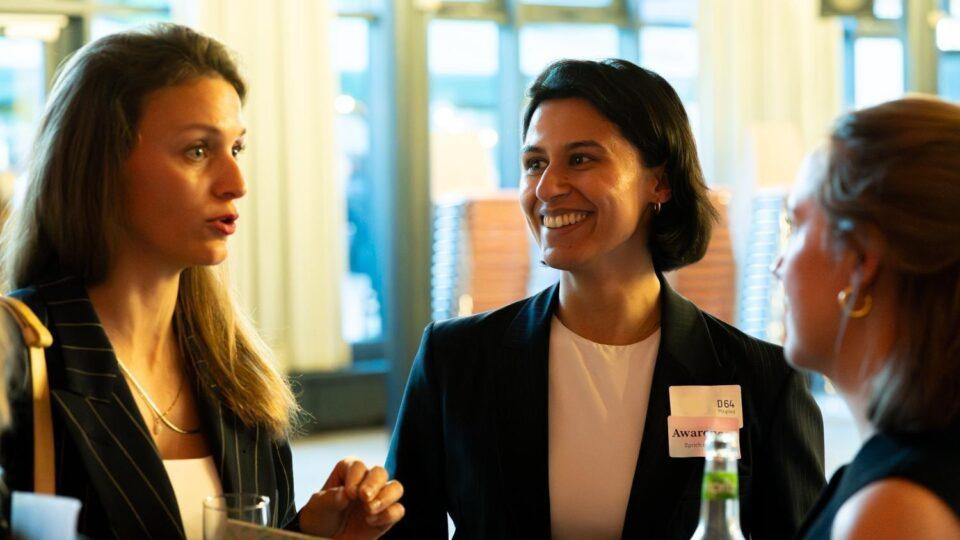 Foto von drei Personen bei einer Veranstaltung in einem hellen Raum mit großen Fenstern im Hintergrund. Sie tragen Businesskleidung und unterhalten sich miteinander.