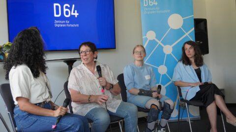 Vier Frauen sitzen nebeneinander auf Stühlen und halten Mikrofone. Sie sind in einer Diskussionssituation. Die zweite Person von links spricht gerade. Im Hintergrund links ein großer Bildschirm mit blauem Hintergrund und dem Text „D64 / Zentrum für Digitalen Fortschritt" in weiß. Rechts dahinter ein großes Poster mit hellblauem Hintergrund, oben „D64 / Zentrum für Digitalen Fortschritt", darunter eine Netzwerk-Grafik mit weißen Kreisen und Verbindungslinien. Links im Vordergrund eine Blume.